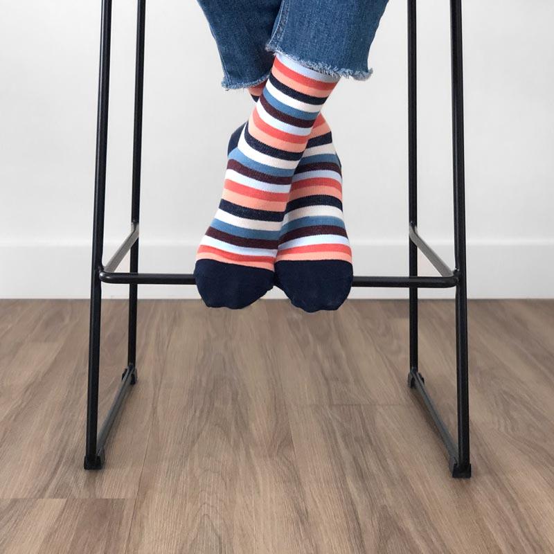 Woman's feet on bar stool wearing stripe patterned compression sock in shades of coral, navy and cream